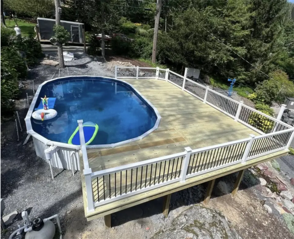 Above ground pool with large wooden deck and white and black railing surrounded by trees.