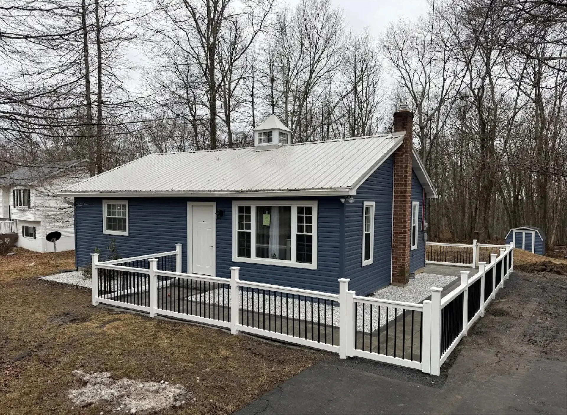 Blue cottage with white trim, fenced patio, and wooded backyard.