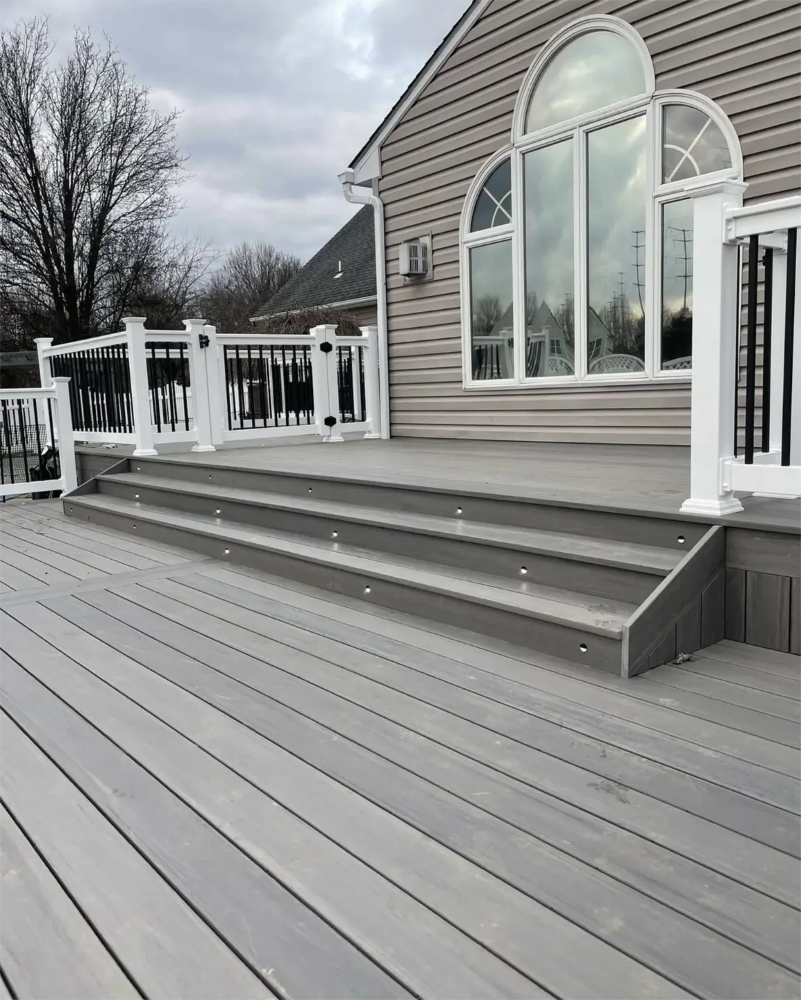Gray composite deck with wide stairs and white railings beside large arched windows.