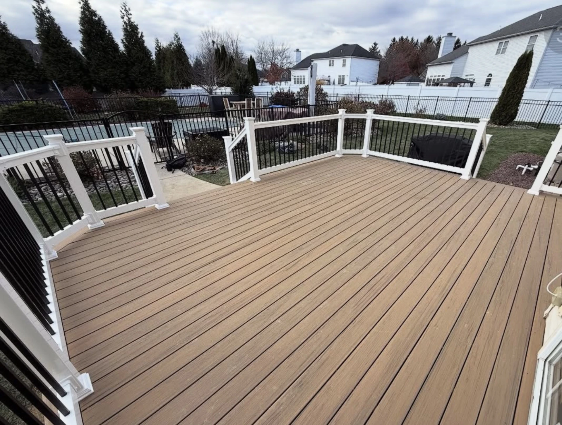 Large tan composite backyard deck with black and white railing overlooking a fenced suburban yard and patio area.