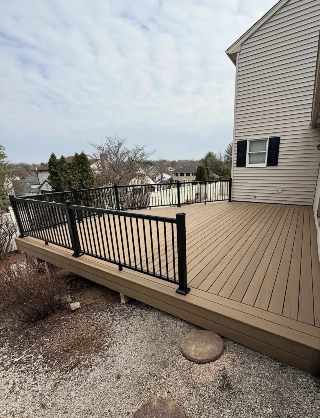 Large tan composite deck with black metal railing beside beige house.