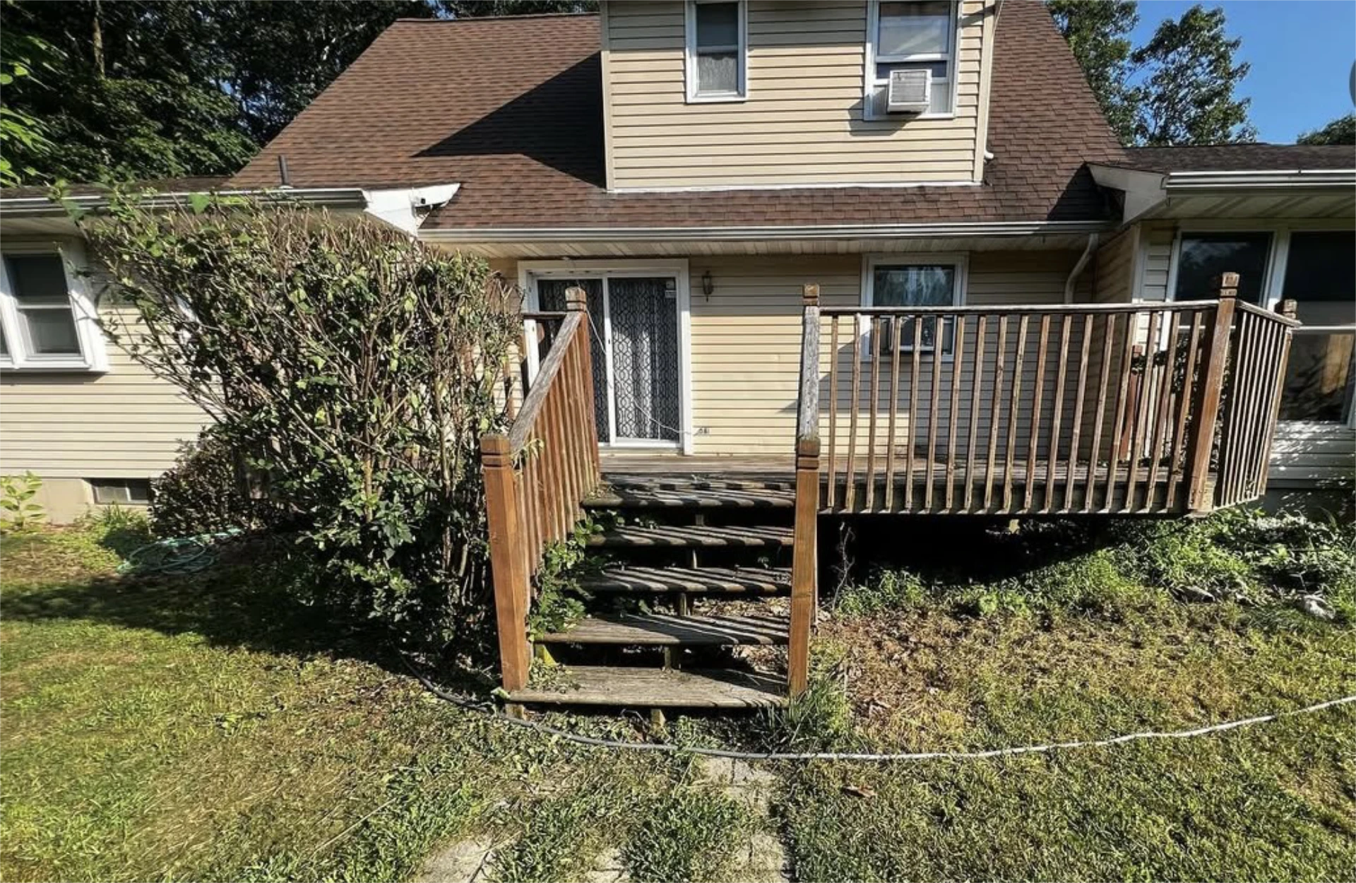 Old wooden backyard deck with weathered railings, worn stairs, and overgrown landscaping behind house.
