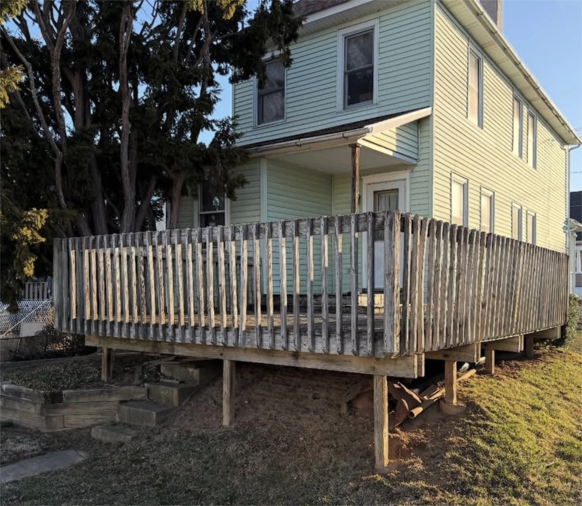 Weathered wooden deck beside pale green house on sloped yard.